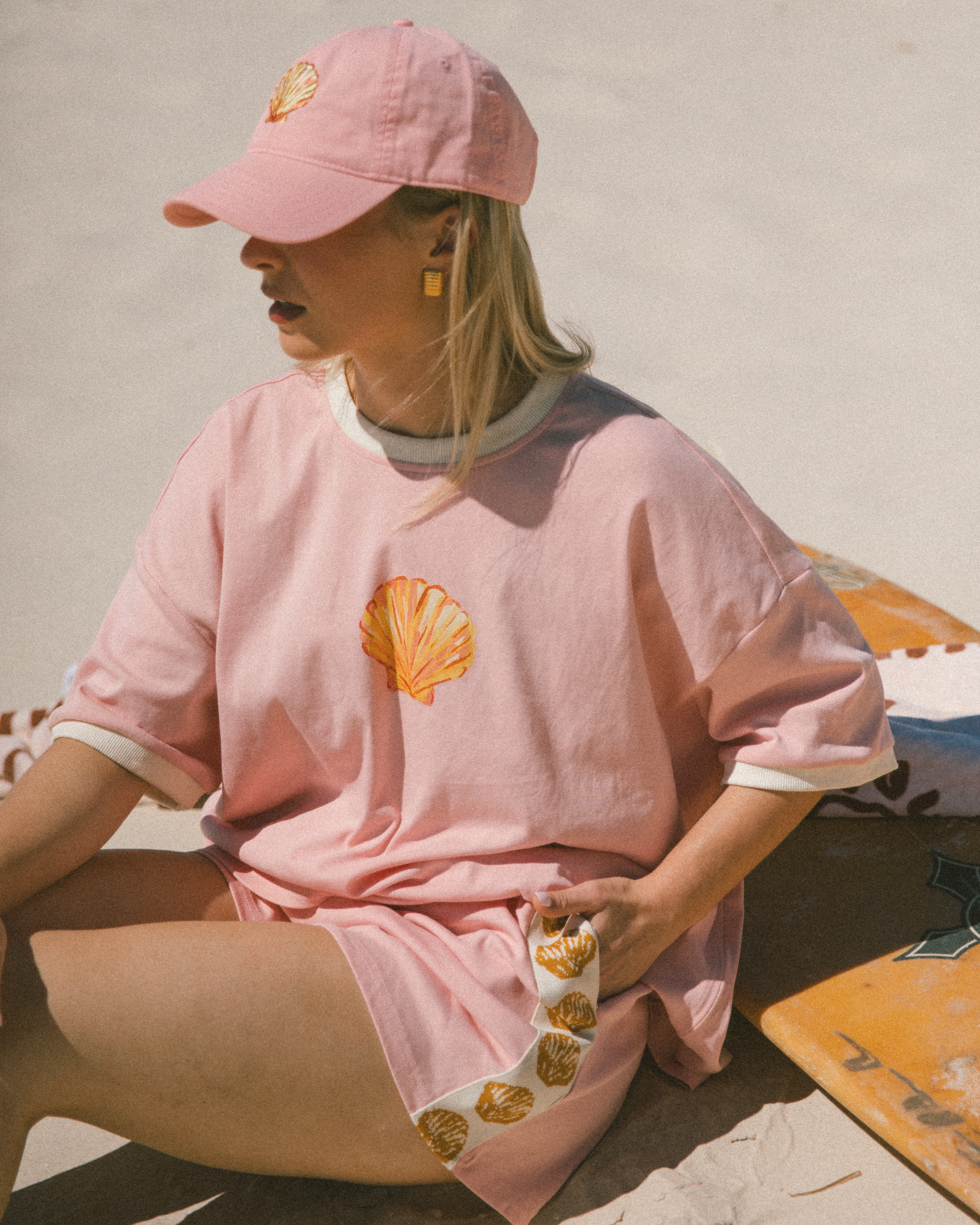 Woman wearing matching pink & gold shell loungewear set and pink cap, sitting on the beach.
