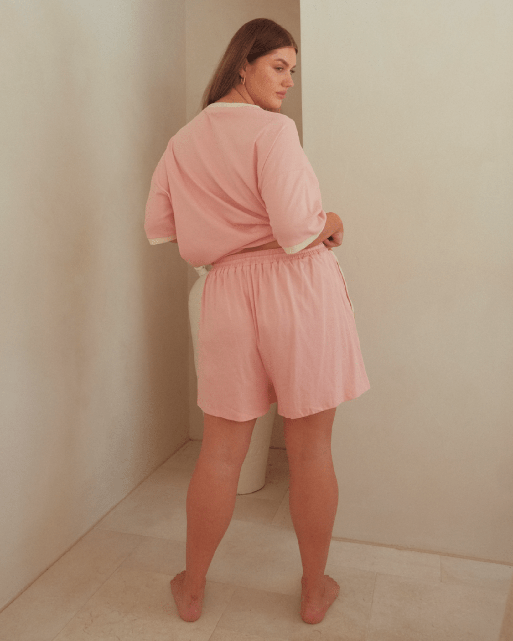 Woman wearing matching co-ord lounge set in pink with cream detailing. Woman is showing the elastic waistband of the high-waisted shorts.