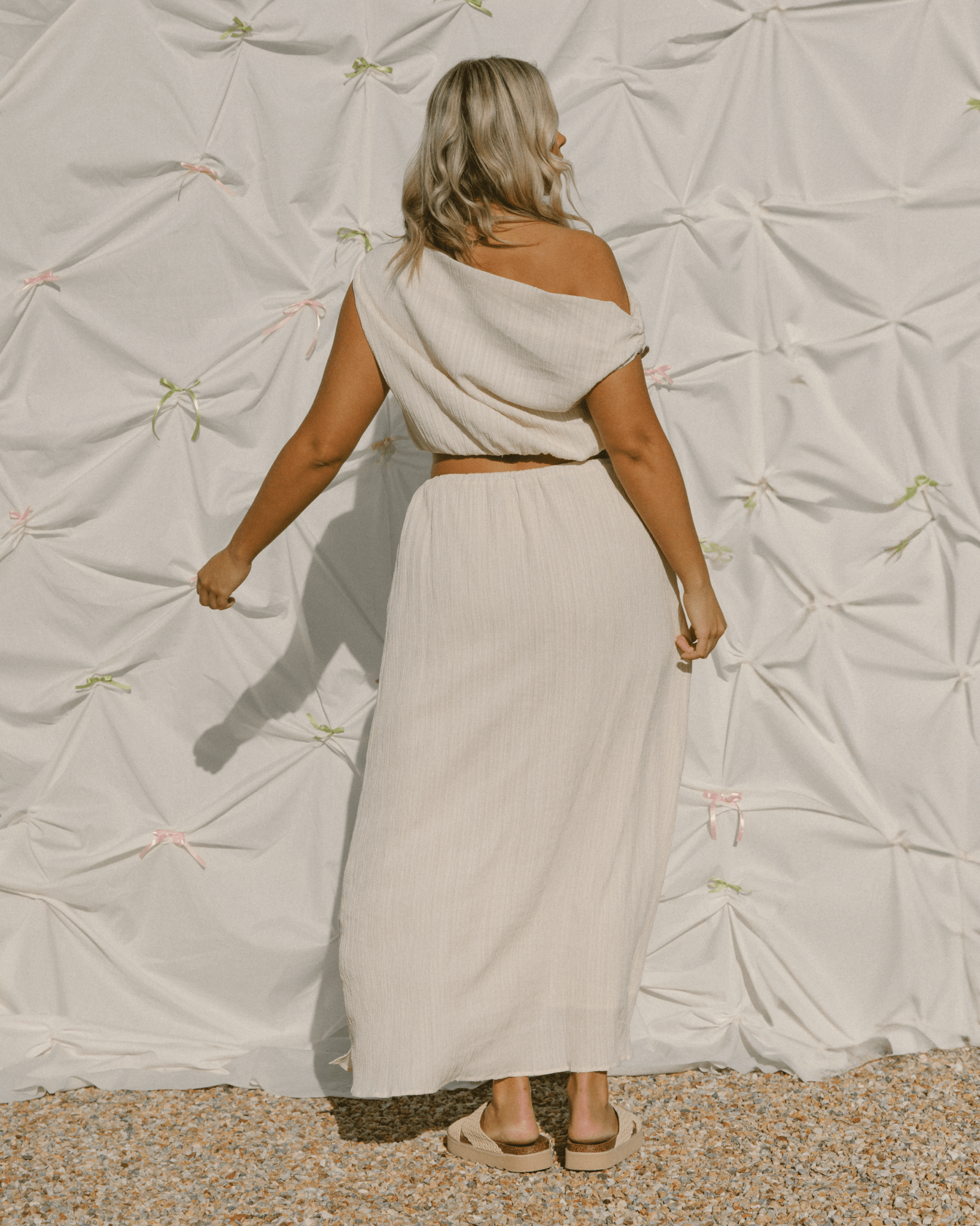 Woman wearing a beige outfit standing against a textured white wall.