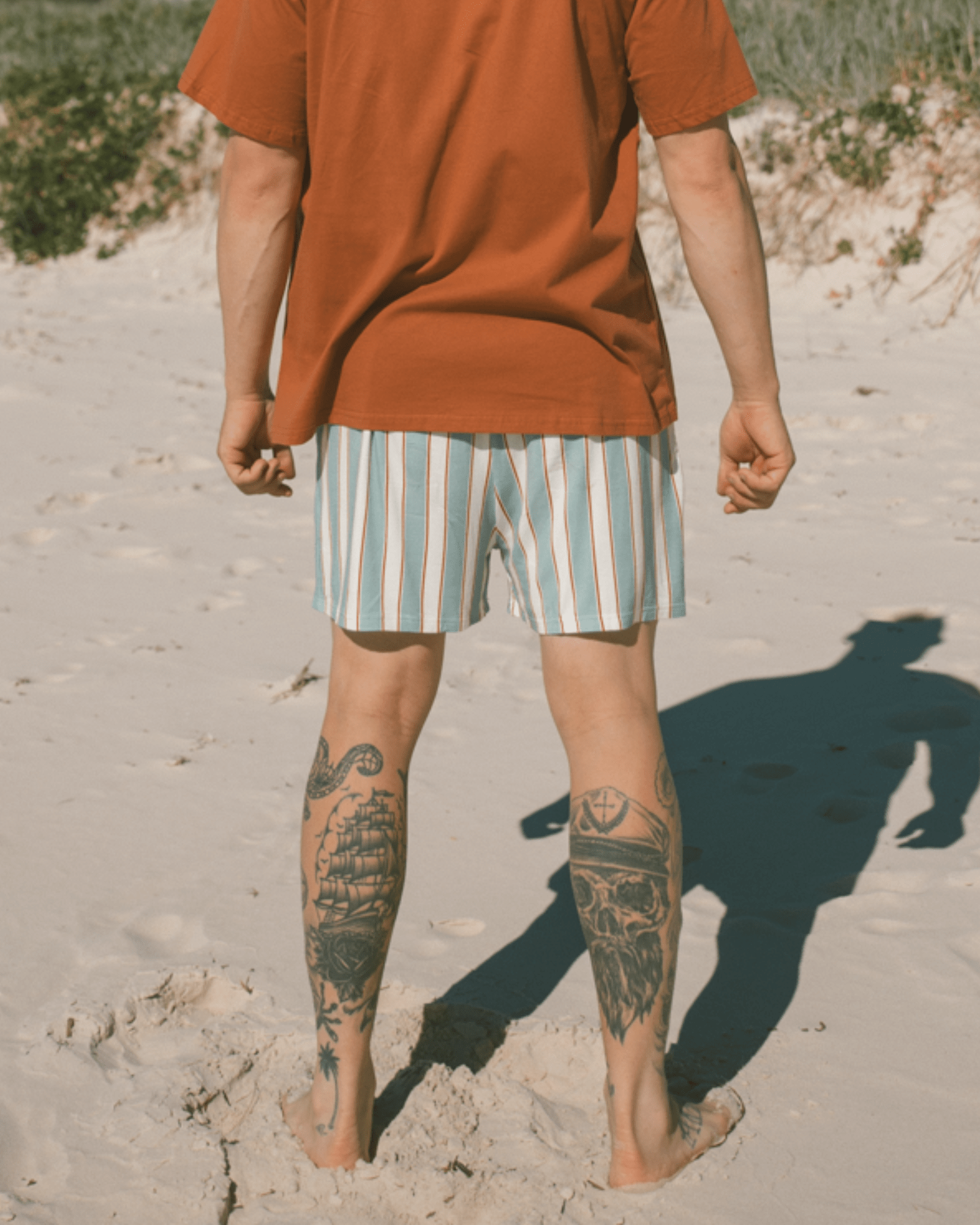 Person wearing a rust-colored shirt and striped shorts on a sandy beach.