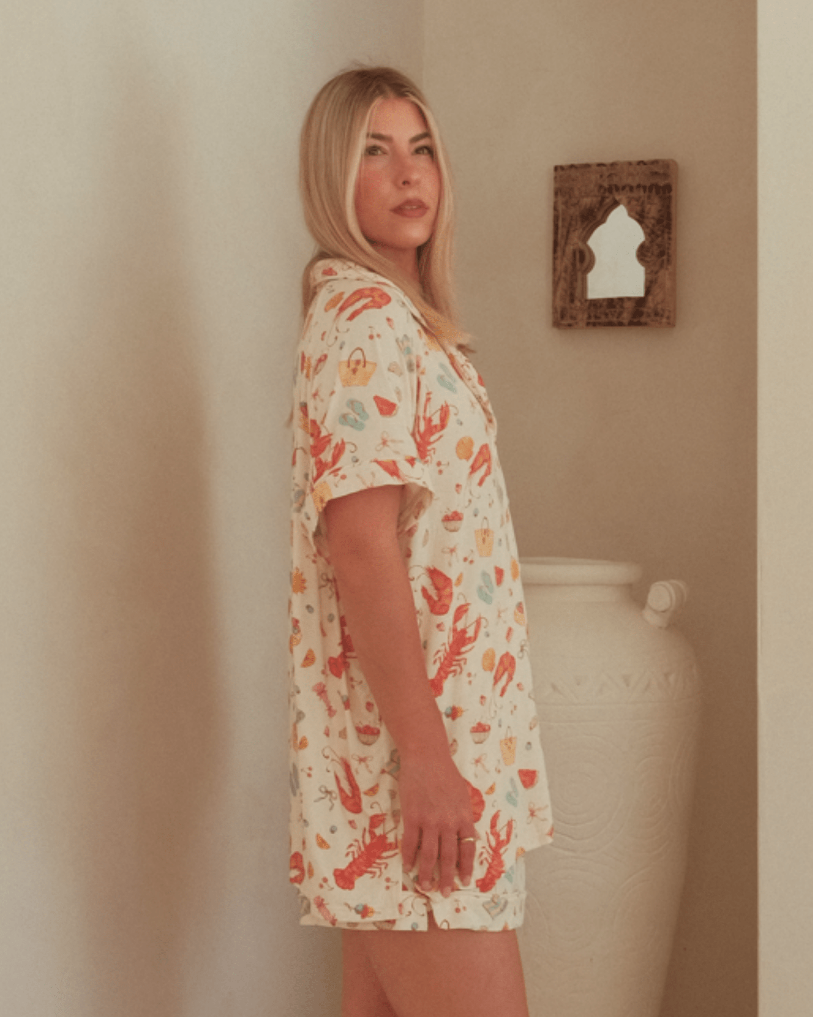 Woman wearing a pattern matching pj's standing against a beige wall with a decorative mirror.