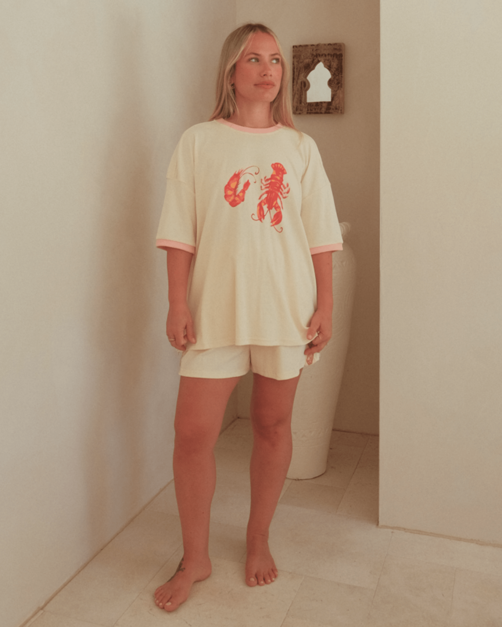 Woman wearing a cream-colored outfit with a red lobster design in a minimal indoor setting.