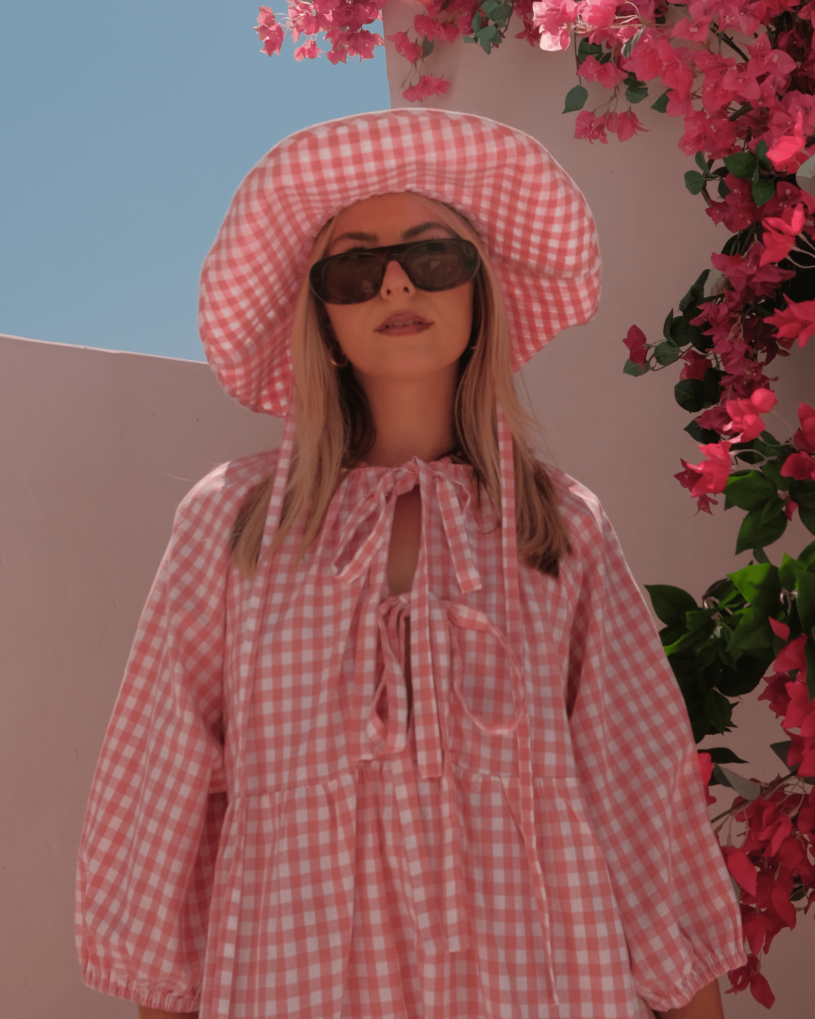 Person wearing a pink checkered dress and hat with sunglasses, standing against a floral background.
