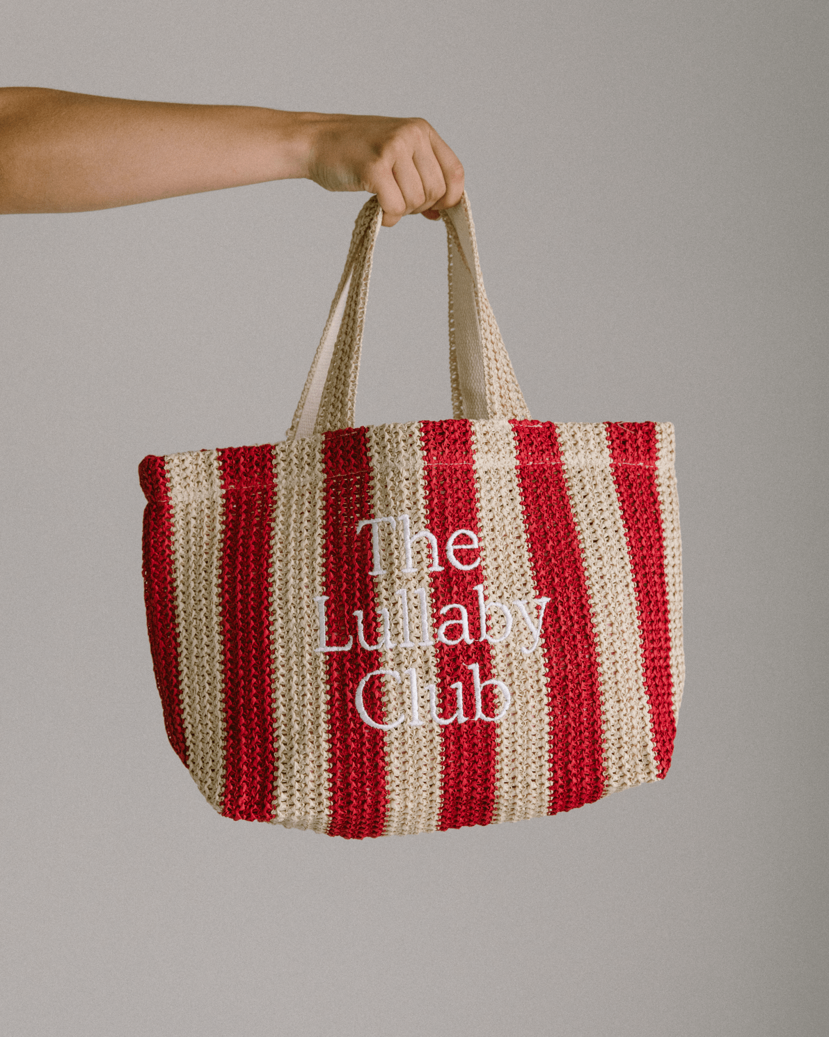 Women holding a woven red and beige striped tote bag with 'The Lullaby Club' embroidered on the front.