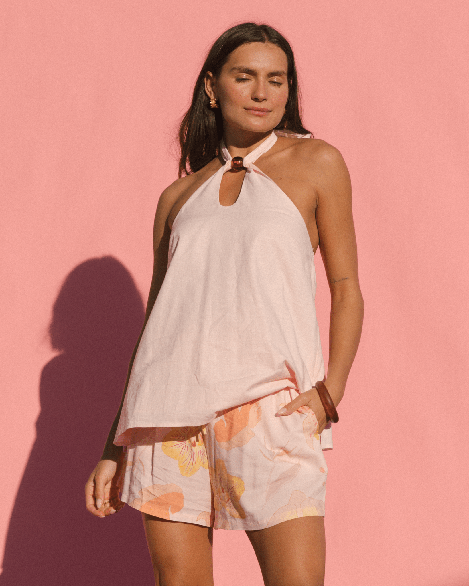 Woman wearing a pale pink halter top and floral shorts against a pink background