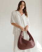 Woman holding a woven wine coloured handbag against a plain background