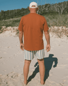 Man walking on a sandy beach wearing an orange shirt and striped shorts.