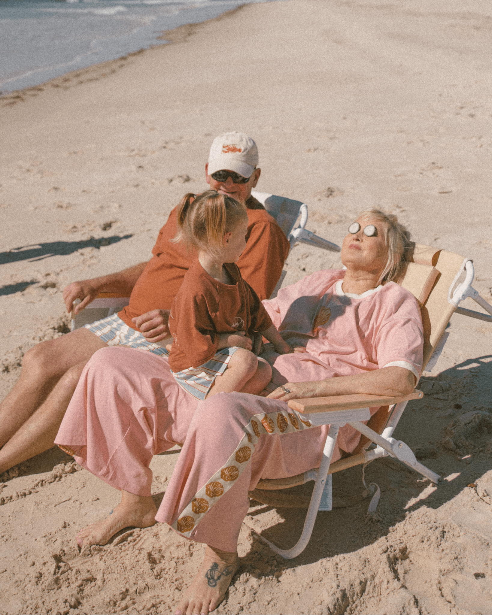 Two people sitting on a beach, one in a pink lounge set and sunglasses, the other in an orange shirt and cap.
