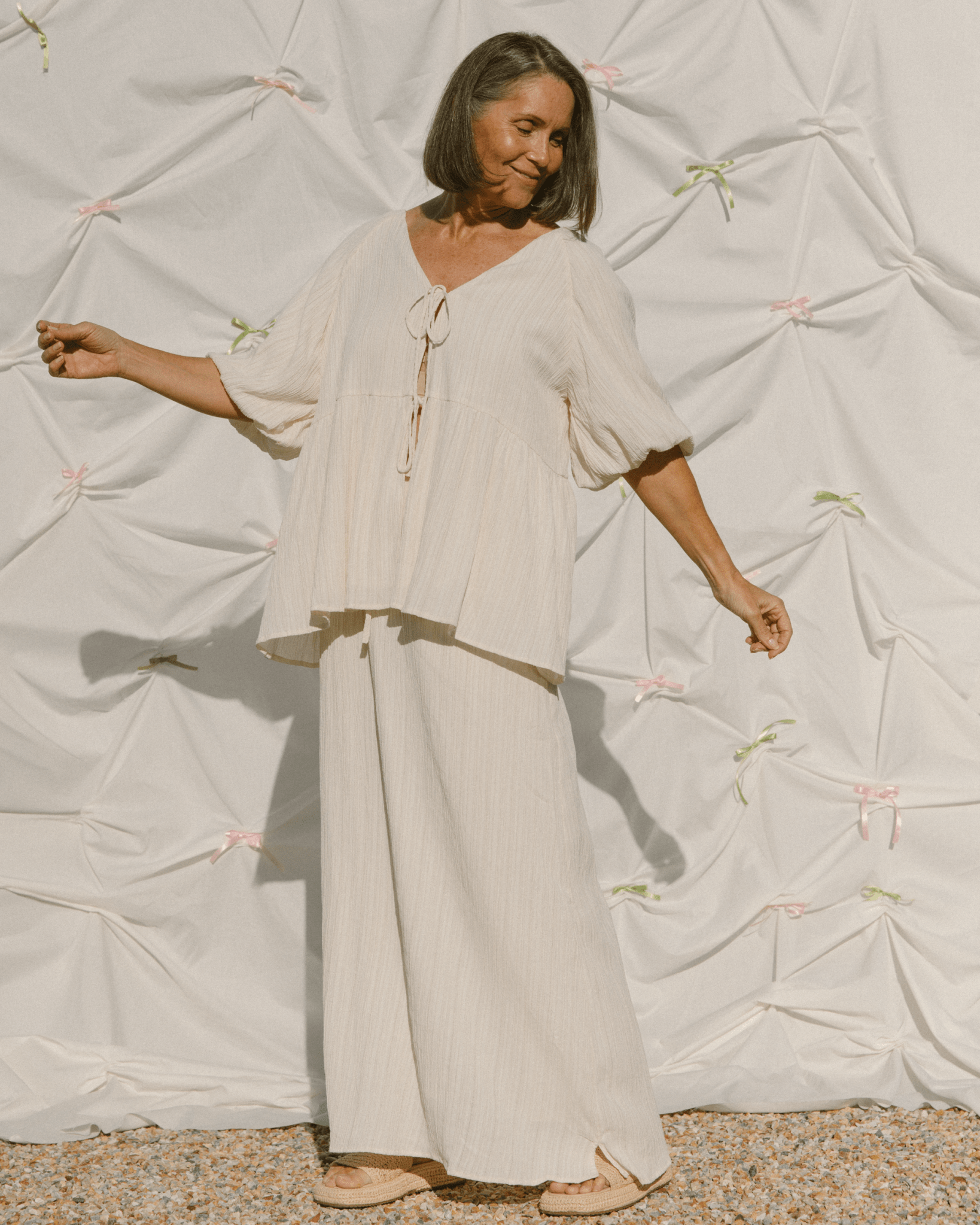 Woman in a flowing beige outfit standing against a textured white wall.