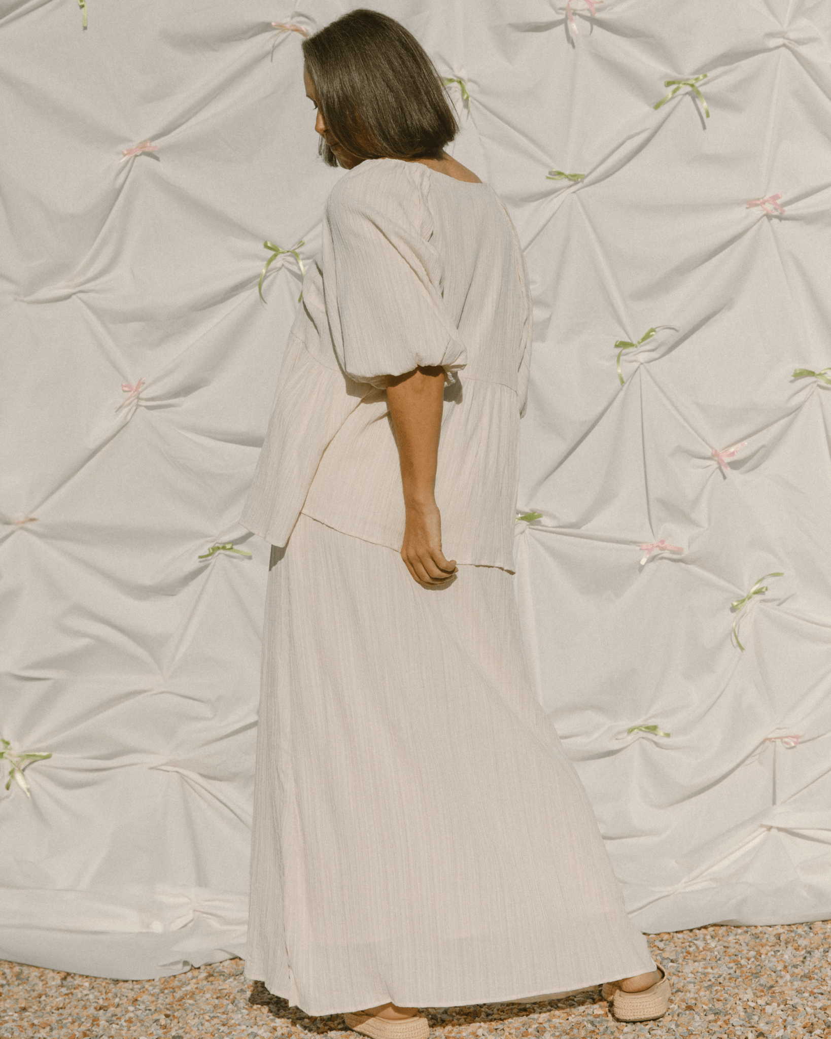 Woman wearing a long, light-colored dress standing against a textured wall.