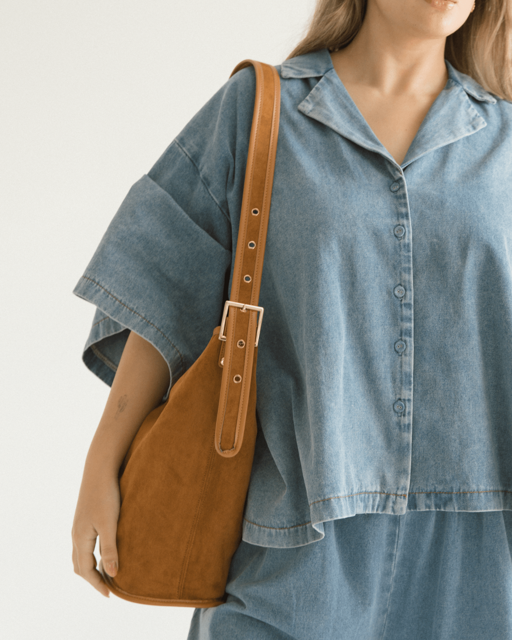 Person wearing a blue denim shirt with a brown leather shoulder bag on a plain background