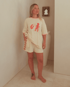 Woman wearing a cream-colored outfit with a red lobster design in a minimal indoor setting.