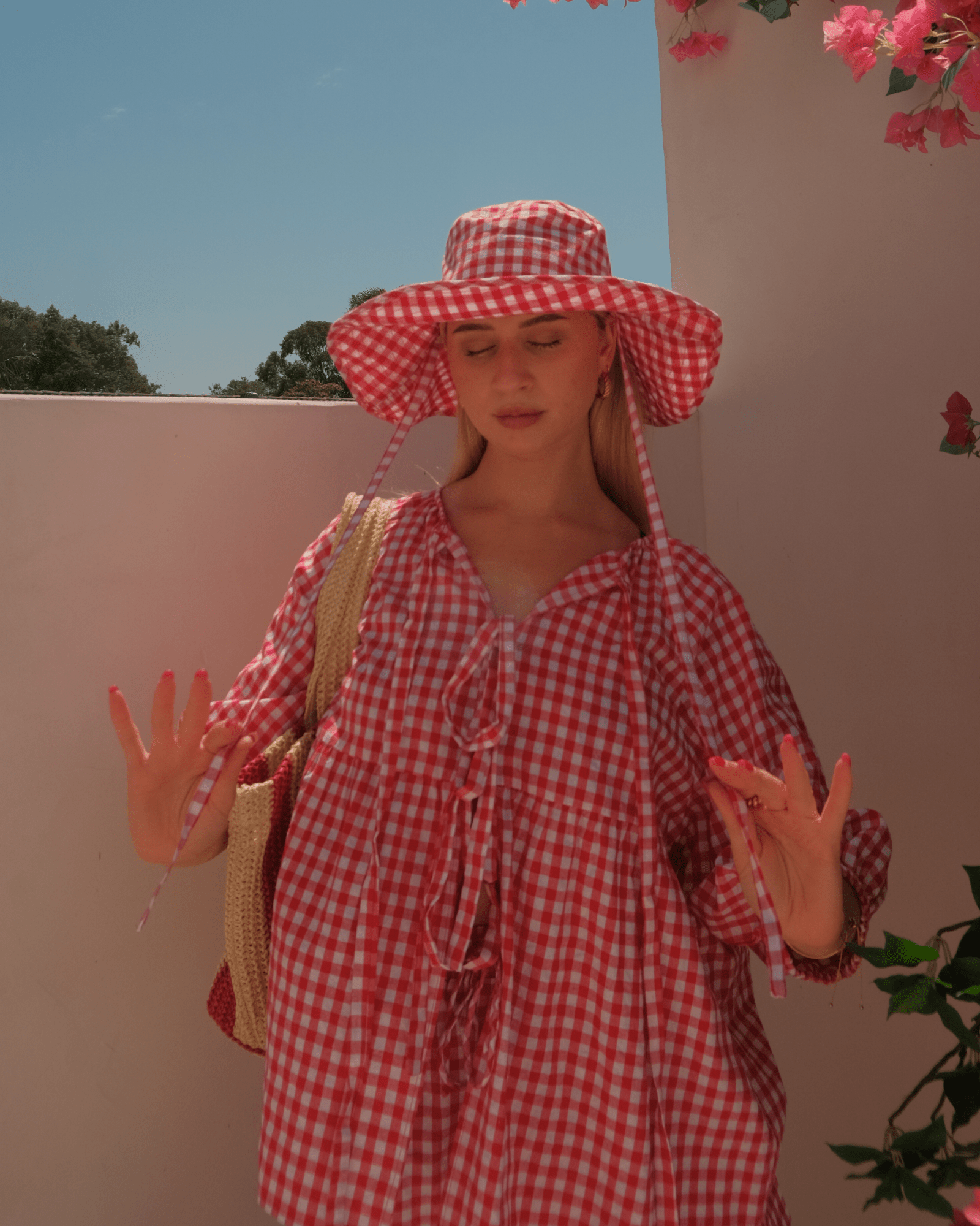 Woman wearing a red gingham dress and matching hat, standing against a light pink wall with greenery.
