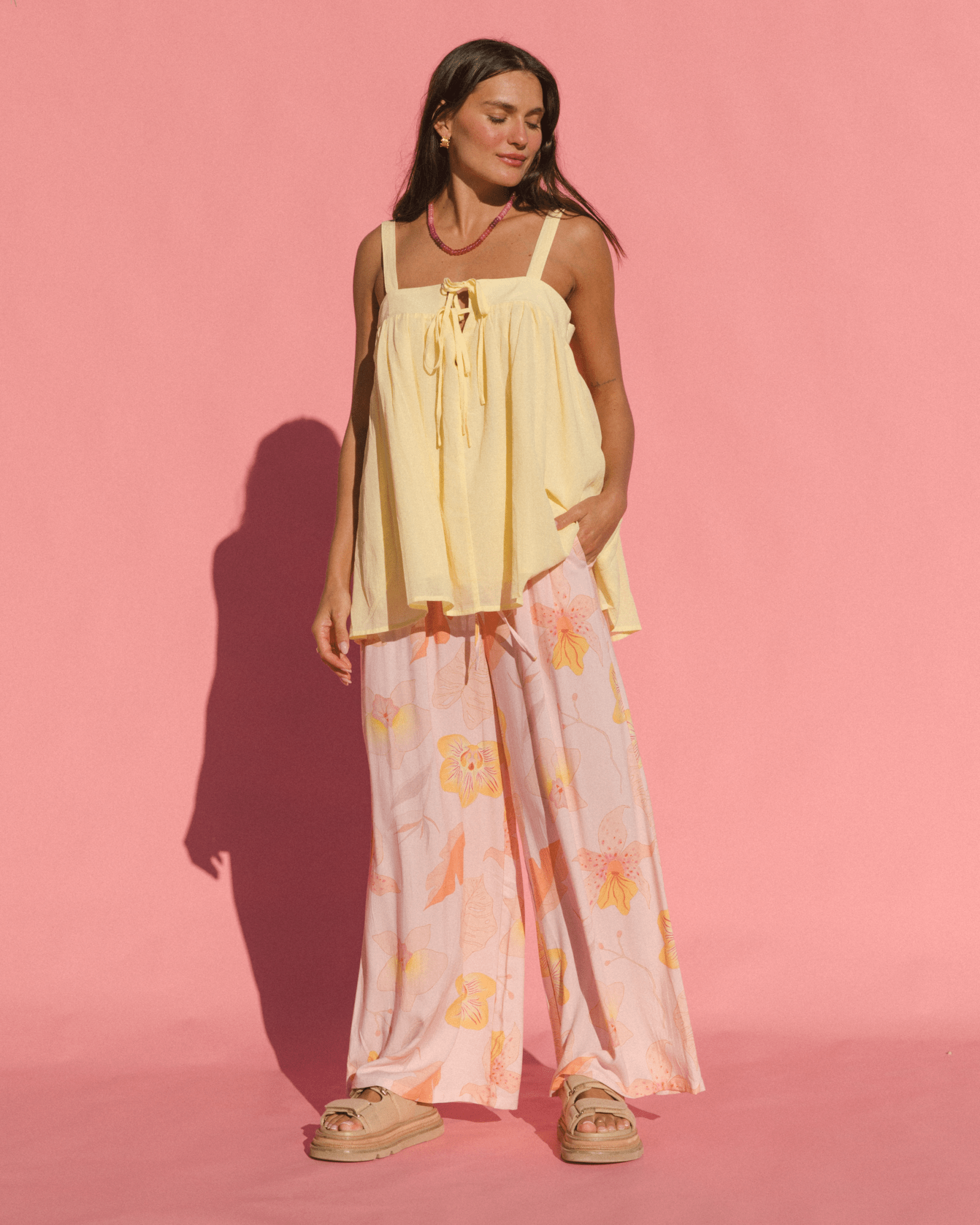 Woman wearing a flowy yellow cotton top and light pink  floral pants against a pink background
